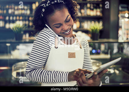 Smiling young African entrepreneur standing in her cafe taking cellphone reservations and using a digital tablet Stock Photo