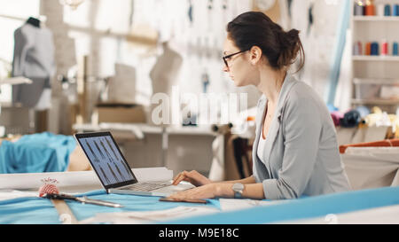 Beautiful Woman fashion, designer,  Sitting at Her Desk Looking at Sketches on a Laptop, Taking Notes. Her Studio is Sunny, Colorful Fabrics Stock Photo
