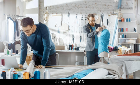Two Male Fashion Designers Working for Their New Collection. Using Laptop and Working With a Dummy. Studio is Sunny. Colorful Fabrics, Clothes Hanging Stock Photo