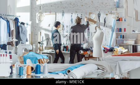 Female and Male fashion, designer,  Choosing and Pinning  to the Wall Clothing Sketches for Their New Collection. Studio is Sunny. Colorful Fabrics Stock Photo