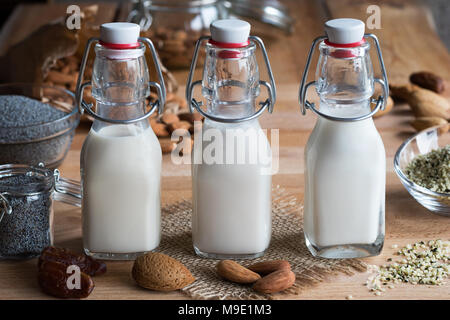 Three bottles of vegan plant milk - almond milk, poppy seed milk and hemp seed milk, with almonds, poppy seeds, hemp seeds and dates in the background Stock Photo