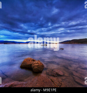 Cloudy sunrise over Jindabyne lake in NSW snowy mountains region of Australia. Clear water of mountain lake shows rocky botton and reflects sunlight. Stock Photo