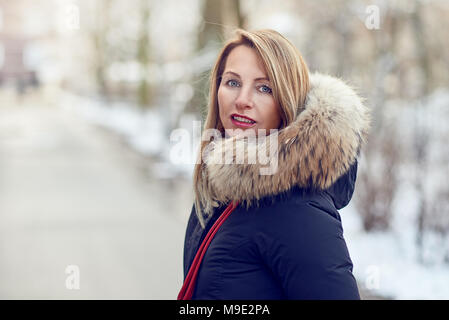 Attractive blond woman wearing a warm jacket with fur lined hood standing outdoors in winter on a snowy road turning to look back at the camera Stock Photo