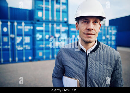 Port manager holding a clipboard and wearing a hardhat while standing alone on a large commercial shipping dock Stock Photo