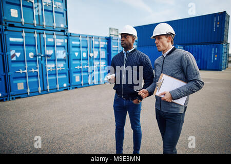 Two engineers wearing hardhats and talking together while standing in a freight yard full of shipping containers Stock Photo