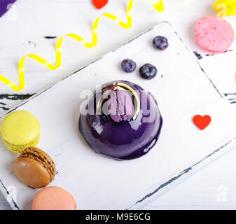 round cake with lilac fondant and decor on a white board, vintage toning, beside colored pastry macarons, top view Stock Photo