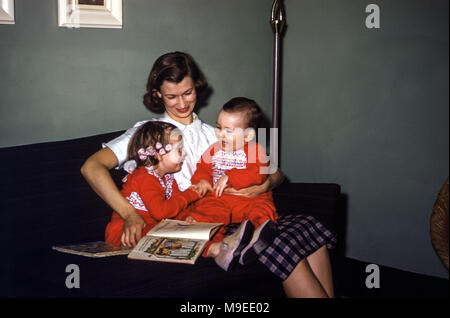 Two young children at bedtime wearing matching pyjamas sitting on their mother's lap on a sofa in a family living room with story books in the 1950s, USA. The little girl is laughing and has hair curlers in her hair. The baby boy is laughing at his sister Stock Photo