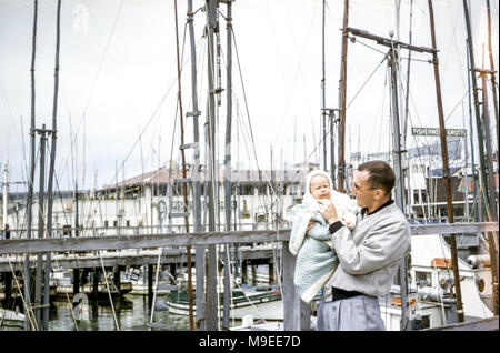 Young man holding a 4 to 5 month old baby girl wrapped in a shawl standing next to masts of boats, Fisherman's Wharf, San Francisco Bay, California, USA in the 1950s Stock Photo
