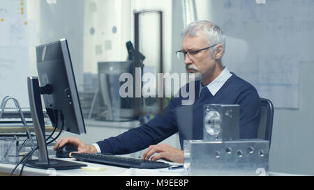Engineering Bureau. Chief Engineer Working at His Desk, Some Technical Components Lying on His Desk. His Assistant Works at His Desk in the Background Stock Photo