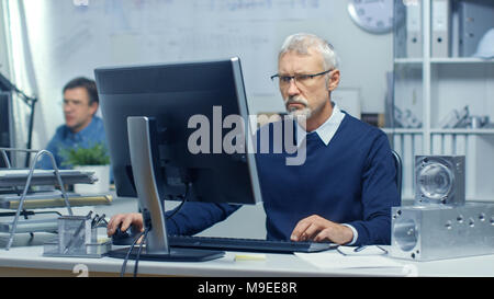 Busy Engineer Bureau. Chief Engineer Working at His Computer, Some Technical Components Lying on His Desk. His Assistant Works at His Desk in the Back Stock Photo