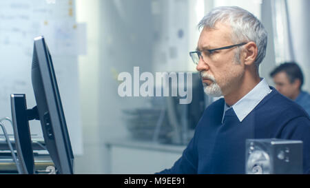 Close-up Shot of a Chief Engineer Working at His Desk, Some Technical Components Lying on His Desk. His Assistant Works at His Desk in the Background. Stock Photo