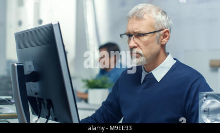 Engineering Bureau. Chief Engineer Working at His Desk, Some Technical Components Lying on His Desk. His Assistant Works at His Desk in the Background Stock Photo