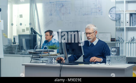 Engineering Bureau. Engineer Working at His Desk, Some Technical Components Lying on His Desk. His Assistant Works at His Desk in the Background. Stock Photo