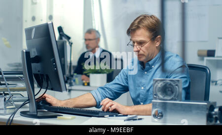 Busy Engineering Bureau. Engineer Working at His Desk, Some Technical Components Lying on His Desk. His Assistant Works at His Desk in the Background. Stock Photo