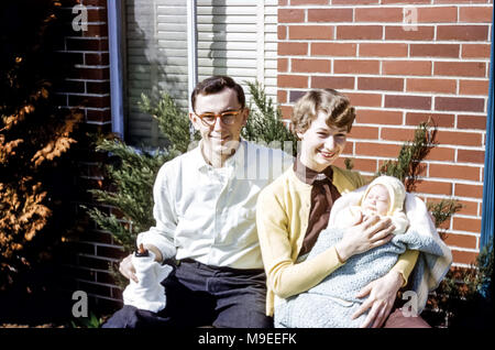 Young couple sitting in sunshine outdoors smiling. The young woman is holding  a newborn baby girl wrapped in a shawl and the young man is holding a baby bottle, USA in 1950s Stock Photo