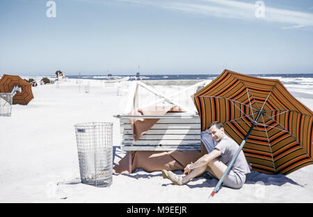 Young man sitting on a white sandy beach next to Atlantic Ocean under a large sun umbrella on a windy day, New York, USA in 1950s Stock Photo