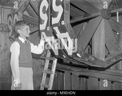 Boy inside an old wooden cricket scoreboard hut, dwarfed by the massive score 'wheels', c. 1950. The wheels were turned by hand by the scorers inside to display the correct number in a window at the front of the hut to the players and spectators at the sports venue. These days most sport scoreboards are electronic and digital displays - a vintage nineteen fifties photograph Stock Photo