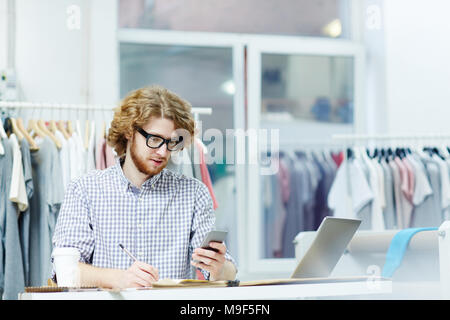 Young businessman sitting at the table looking at his phone and making notes Stock Photo