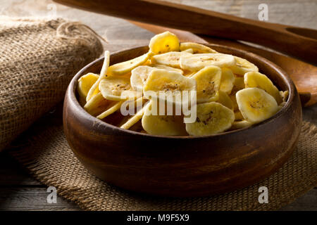 Heap of dried banana chips snack in wooden bowl on burlap on dark wooden table Stock Photo
