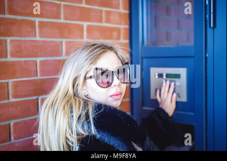 young blond woman in a fur coat, dials the code on the doorphone panel, presses  buttons with her fingers to get inside house Stock Photo
