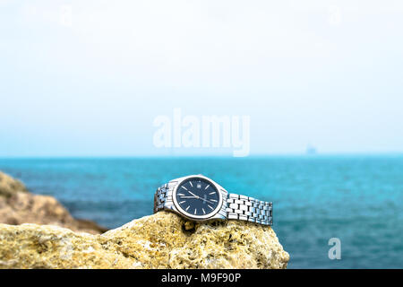 an exotic stainless silver watch kept on the brownish rocks at the beach and greenish sea water in the background cloudy day Stock Photo