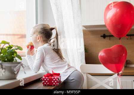 Cute preschooler girl celebrating 6th birthday. Girl eating her birthday cupcake in the kitchen, surrounded by balloons. Stock Photo