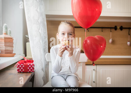 Cute preschooler girl celebrating 6th birthday. Girl eating her birthday cupcake in the kitchen, surrounded by balloons. Stock Photo