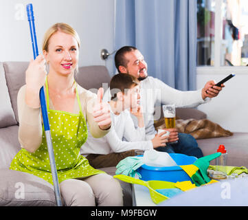 https://l450v.alamy.com/450v/m9fbgw/smiling-woman-dressed-for-cleaning-sitting-on-sofa-while-husband-with-son-relaxing-m9fbgw.jpg