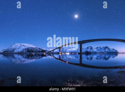 Amazing bridge and starry sky with beautiful reflection in water. Night landscape with bridge, snowy mountains, blue sky with moon and bright stars re Stock Photo