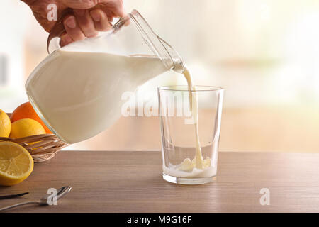 Filling glass of milk with jug for breakfast on brown wooden table in white kitchen. Horizontal composition. Front view Stock Photo