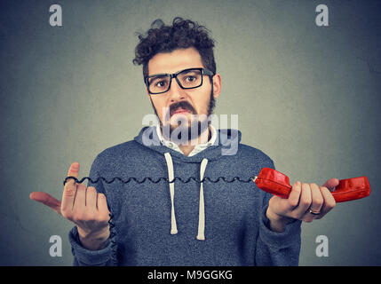 Disgusted young man looking at camera with bewilderment and confusion holding old fashioned red handset. Stock Photo