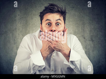 Young shocked man in white shirt covering mouth holding scream while looking at camera with amazed face expression. Stock Photo