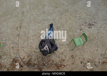 Man sitting on chair in city park wearing coat and hood, sleeping, with hands in pockets - aerial view Stock Photo