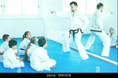 Cheerful young male coach explaining new maneuvers to children in karate class. Focus on the boy Stock Photo