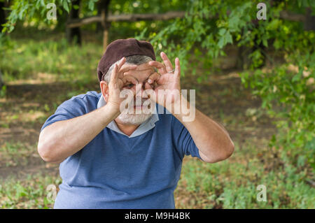 Portrait of queer senior making gesture like looking through binocular while resting in summer park Stock Photo