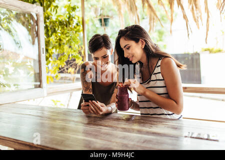 Two smiling women sitting at a restaurant looking at mobile phone. Woman showing mobile phone while another woman holds a smoothie jar in hand. Stock Photo