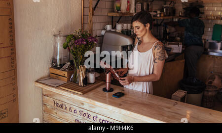 Woman standing at the billing counter of her coffee shop. Coffee shop owner operating the billing machine while a worker prepares coffee in the backgr Stock Photo