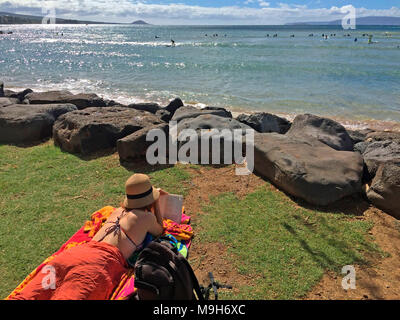 A youngwoman reclining, relaxing, and reading in Kalama Park, overlooking the ocean in South Kihei, Maui, Hawaii, USA Stock Photo