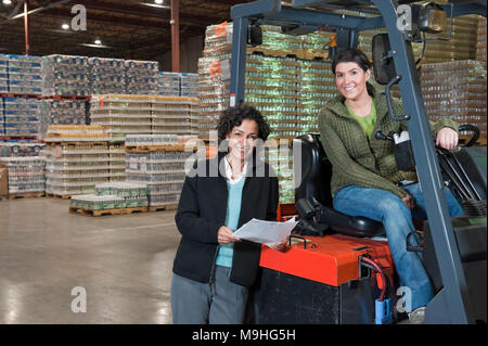 Portrait of two multi-ethnic female warehouse workers on a forklift in a large warehouse full of pallets of flavored water in cans. Stock Photo