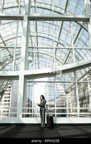 Woman in a business suit standing with her pull along suitcase, using a smart phone, under a glass atrium roof. Business travel. Stock Photo