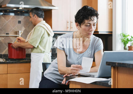 A man cooking supper at the hob, and a woman using a laptop and checking paperwork in the kitchen. Stock Photo
