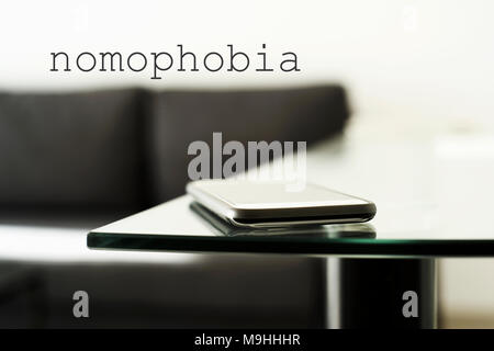 closeup of a smpartphone on a glass table in a modern living room or a lobby, and the text nomophobia, for the phobia of leaving home without the mobi Stock Photo