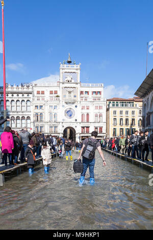 Tourist with luggage wading trough Acqua Alta flooding in Piazza San Marco (St Marks Square), Venice, Italy with lines of people on passerelles Stock Photo