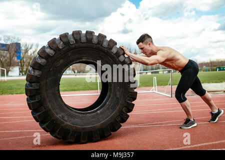 Young muscular man flipping a huge tire for training muscles at the tartan track on the stadium. Stock Photo
