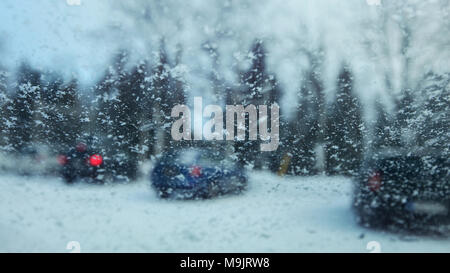 View from inside car to snow flakes on windshield with blurred cars in parking lot in background. Winter weather driving conditions. Stock Photo