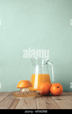 Whole oranges and a half orange on manual squeezer with a glass jug of orange juice behind on a wood plank table with pale turquoise background. Filte Stock Photo