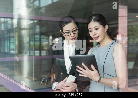 Asian supervisor showing their product to client standing aside the glass door. Using the tablet inside the modern and advanced company. Stock Photo