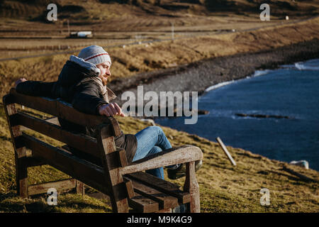 Man relaxing on a wooden bench by the water on Isle of Sky, Scotland, on a sunny spring day. Stock Photo