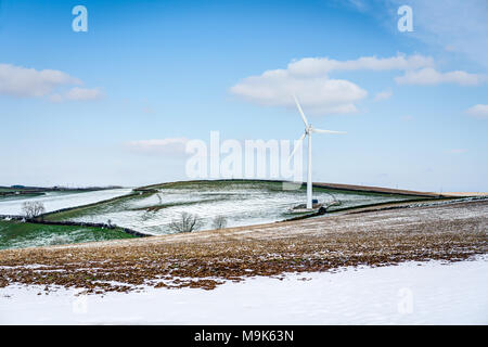 A photograph of a large wind turbine standing on Cornish farmland, pictured in winter with the surrounding fields showing melting snow, grass and soil Stock Photo