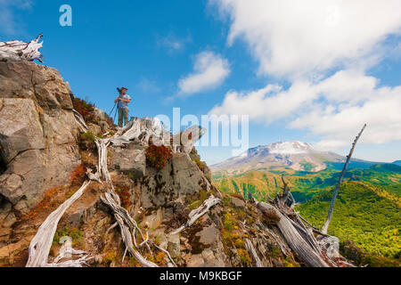 A hiker/photographer looks away from his tripod and camera, on top of Castle Peak with with Mt. St. Helens in the background in Gifford Pinchot Nation Stock Photo